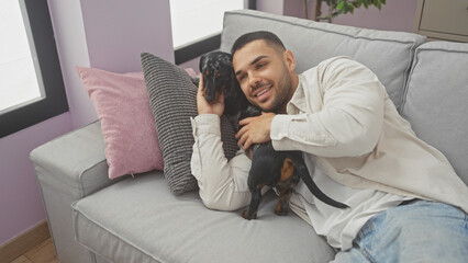 Poster - A young hispanic man smiles while cuddling with a dachshund on a couch in a cozy living room.