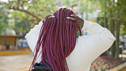 Sticker - Woman with braids in a white sweater posing outdoors surrounded by greenery in a city park.