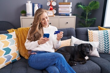 Wall Mural - Young blonde woman using smartphone and credit card sitting on sofa with dog at home
