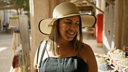 Wall Mural - A smiling woman in a straw hat enjoys exploring a traditional market in dubai, surrounded by spices and sunlight.