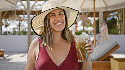 Wall Mural - A smiling woman in a sunhat enjoys a mojito at a luxury beach resort, epitomizing relaxation and leisure.