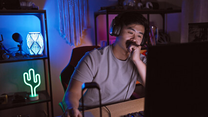 A young asian man, wearing headphones, pensive in a dark gaming room with neon lights