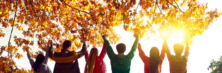 Group of happy friends standing with raised hands at sunset. Rare view of young people holding hands together looking at sunset in autumn park. Friendship, teamwork, excitement concept