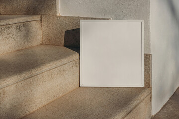 Closeup of empty square white thin picture frame leaning against wall in sunlight. Outdoor sandstone stairs background, shadows overlay. Poster mockup for art display. Minimal summer design. No people