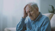 © Cadengo - An elderly man sits on the sofa, holding his head in pain and thinking about loneliness