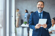 © Dragana Gordic - Smiling mid aged business man wearing suit standing inside office holding digital tablet. Mature businessman professional holding fintech device looking away thinking or new business ideas solutions