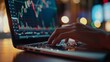 © Plaifah - Close-up of hands typing on a laptop keyboard with a stock chart visible on the screen.