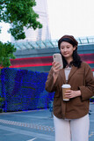 Fototapeta  - Stylish Woman Using Phone by Modern Cityscape