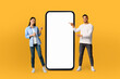 © Prostock-studio - The scene features a young multiethnic couple, both grinning, positioned next to a life-sized smartphone mockup on a seamless yellow background.