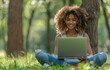 © yevgeniya131988 - Young woman sits under a tree in a summer park, smiling while using a laptop, wearing casual attire