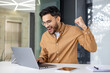 © Tetiana - Happy young muslim man sitting in office at table, looking at laptop screen and enjoying achievement and success, making victory gesture with hand yes