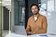 © Tetiana - Portrait of a young Indian man sitting in the office at the desk wearing headphones, working on a laptop and looking seriously into the camera