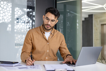 A young Muslim man is working concentratedly in the office at the table with a laptop, making notes of data and documents