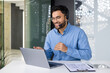 © Tetiana - A young Muslim man wearing a headset and glasses is sitting in the office in front of a laptop and talking online on a video call, smiling and gesturing with his hands