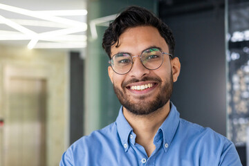Close-up portrait of a young Indian man standing in glasses and blue shirt smiling in a modern office and looking at the camera