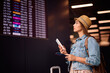 © Drazen - Young woman using cell phone while checking flights departure board.