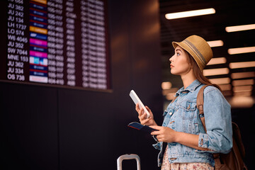 Wall Mural - Young woman using cell phone while checking flights departure board.