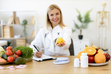 Portrait of smiling female nutritionist doctor sitting at table with vegetables and fruits in clinic. Positive mature Caucasian female weighing lemon while writing healthy food recipe on clipboard.