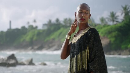 Wall Mural - Outrageous gay black man in luxury gown poses on scenic ocean beach. Gender fluid ethnic fashion model in long posh dress and accessories looks at camera and touches neck gracefully slow motion