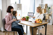 © sofiko14 - Mature Caucasian female doctor shows on tablet graphs of food for healthy eating. Little school girl sitting on mothers lap with visit to woman pediatrician nutritionist.