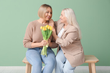 Canvas Print - Adult woman with her mother and bouquet of tulips sitting on bench near green wall. International Women's Day celebration