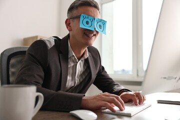 Wall Mural - Man with fake eyes painted on sticky notes working on computer at table in office