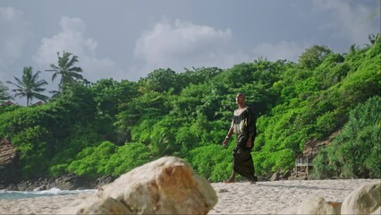 Wall Mural - Gay bipoc man in boutique sparkling glossy dress, jewelry, makeup walks on tropical rocky ocean beach. Homosexual ethnic fashion model in posh gown goes on sandy sea shore against lighthouse, clouds.