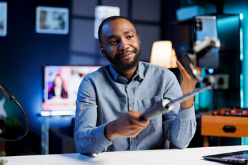 Cheerful entertainer using mobile phone attached to selfie stick to recording himself chatting with online audience. Online star discussing with fans during live broadcast stream from home studio