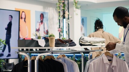 Wall Mural - African american employee working in SH clothing shop, placing simple stylish shirts on racks. Retail assistant formal garments merchandise on hangers in discount fashion store