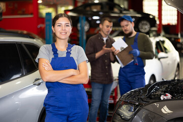 Wall Mural - Portrait of positive girl auto mechanic in uniform against background of cars in a car service center