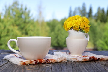 a white cup of hot tea next to a white jug with yellow flowers stands on a wooden desk on the backgr