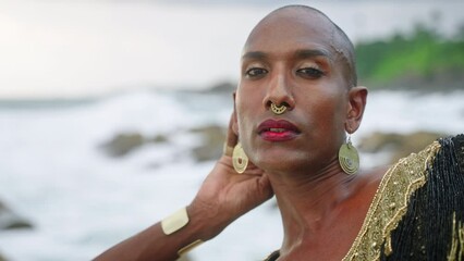 Wall Mural - Gay bipoc man in luxury dress, boutique brass jewelry, makeup poses sitting on tropical rocky ocean beach. Homosexual epatage fashion model in posh gown, nose-ring piercing, earrings, rings with gems.