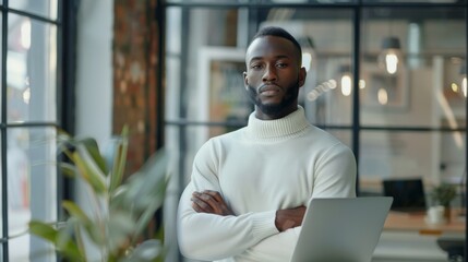 Wall Mural - In an Agency, a handsome Black African American Male stands in a meeting room behind glass walls with a laptop computer. He is wearing a white jumper and working on an app's interface.