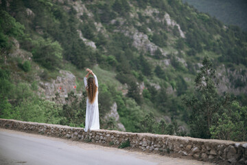 Wall Mural - A woman stands on a stone wall overlooking a mountain. She is wearing a white dress and she is in a contemplative mood.