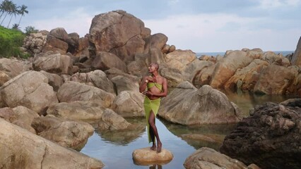 Wall Mural - Gender fluid elegant person of color in beach clothes, jewelry poses on scenic ocean shore rocky pond. Pride extravagant fashion model in open dress in tropical location turns, looks at camera.