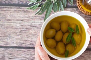 Wall Mural - Hand holding bowl of green olives with leaves in extra virgin olive oil on wooden table. Top view. Copy space. Close-up. Healthy fats, Mediterranean food concept.