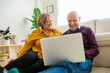 © Jordi Salas - Elderly couple sitting together in a living room looking at a laptop screen. The woman stands behind the man while they both look at the device in a cozy living room.