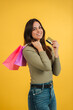 © Jose Calsina - Vertical portrait of a young caucasian customer woman holding shopping bags and a credit card, contemplating whether to buy clothes from an online store. She is against a yellow background purchasing
