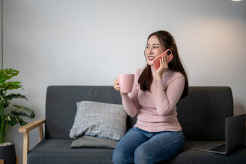 Wall Mural - A woman is sitting on a couch and talking on her cell phone. She is holding a pink mug and a laptop