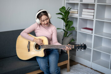 Wall Mural - A woman is playing a guitar in a living room. She is wearing headphones and smiling. The room has a couch, a potted plant, and a bookshelf