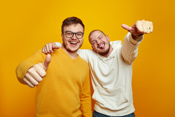 Image of two cheerful handsome guys embracing each other while showing thumbs up, wearing casual clothes, isolated over yellow studio background
