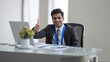 © Aamir - A happy Indian professional man gives a thumbs up while sitting at his desk with a laptop in an office