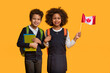 © Prostock-studio - African American boy and girl, both wearing school backpacks and casual clothing, stand side by side with bright smiles. The girl is holding a small Canadian flag, and both are equipped with notebooks