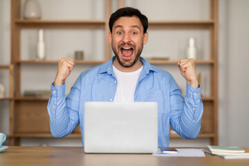 Poster - Excited man is seated at a table with a laptop open in front of him. He appears focused on the screen, possibly working or studying, clenching fists and exclaiming