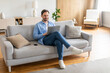© Prostock-studio - A man is sitting comfortably on a couch, focused on his laptop screen. He appears engaged in typing or browsing online content. The room is well-lit, with various items around him.