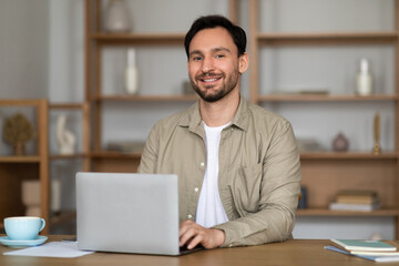 Poster - A man with a cheerful expression is seated at a desk with a laptop computer in front of him. He appears engaged in his work, typing on the keyboard and looking at the screen attentively.