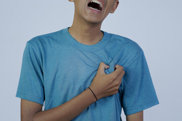 close up portrait of young Asian man in blue shirt moaning in pain while holding his chest, heart disease, isolated in gray background.