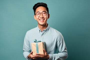 Wall Mural - Portrait of a glad asian man in his 20s holding a gift isolated in plain cyclorama studio wall