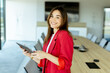 © BGStock72 - Smiling professional woman in red blazer holding tablet in modern conference room during daytime