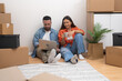 © olga_demina - Couple sits on bare floor among cardboard boxes looking at laptop checking availability of decorations for placing in interior of house in electronic list